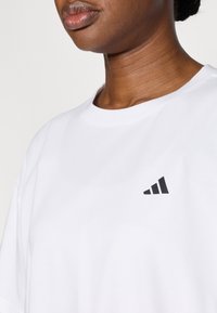 T-shirt en coton blanc avec un col rond et des manches courtes, arborant un petit logo noir sur le côté gauche de la poitrine. Texture lisse.