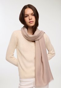 Beżowy sweter z dzianiny w serek, w zestawieniu z jasnobeżowym, teksturowanym szalem owiniętym wokół szyi, z postrzępionymi brzegami.