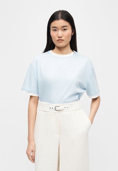 Νεαρή γυναίκα με ίσια σκούρα μαλλιά φοράει ανοιχτό γαλάζιο μπλουζάκι με κοντά μανίκια, στερεωμένο μέσα σε λευκό υφασμάτινο παντελόνι ψηλής μέσης με υφή και ζώνη.