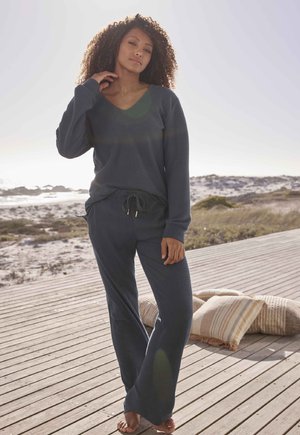 Completo da lounge a coste di colore blu scuro, composto da una maglietta a maniche lunghe con scollatura a V e pantaloni con coulisse dal fit rilassato. Ambiente naturale con cuscini nelle vicinanze.