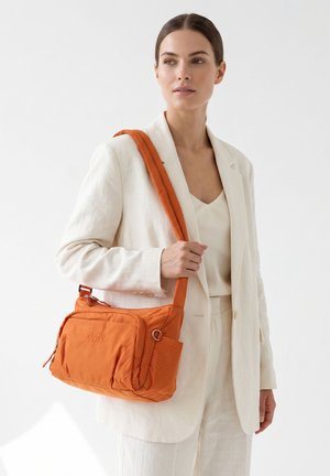 Vrouw gekleed in een gebroken witte pak met een oranje crossbodytas met meerdere ritsvakken en zichtbaar "GMV"-logo.