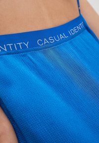 Blaue Textilie mit einem strukturierten Muster, oben mit einem blauen Band versehen, das in weißem Text "CASUAL IDENTITY" trägt.