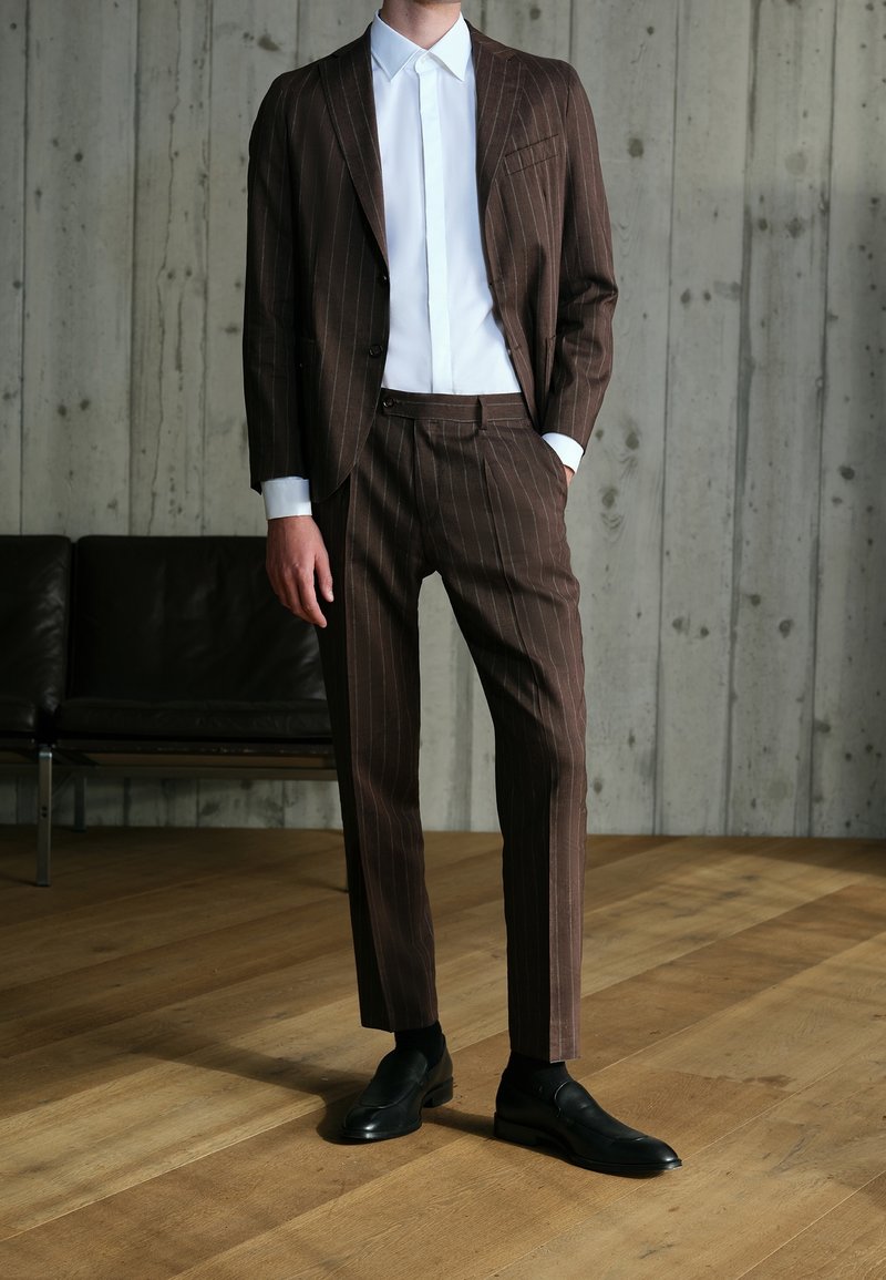 Homme portant un costume marron à fines rayures avec une chemise blanche et des chaussures noires, debout sur un sol en bois devant un mur en béton gris.