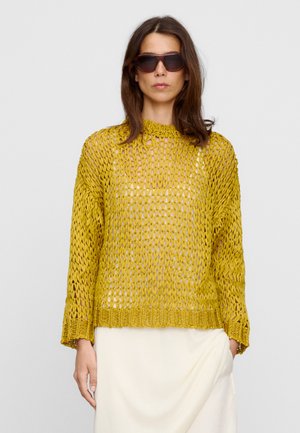 Donna che indossa occhiali da sole scuri, un maglione giallo a maglia larga e una gonna bianca si trova contro uno sfondo chiaro e semplice.