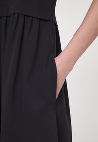 Main reposant dans la poche d'une robe noire plissée sur un fond clair uni.
