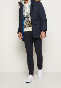 Muž ve námořnické parce s kožušinovou kapucí, potištěné mikině, tmavých kalhotách a bílých teniskách, stojící na jednolitém světlém pozadí.