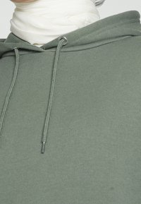 Primer plano de una persona que lleva una sudadera verde salvia con cordones y un cuello de tortuga blanco debajo.