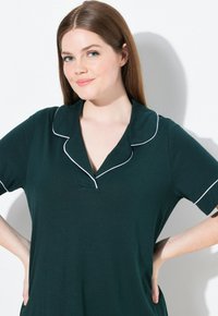 Abito corto verde scuro con maniche corte, colletto a rever, piping bianco e texture morbida. Il design presenta una vestibilità comoda e uno stile informale.