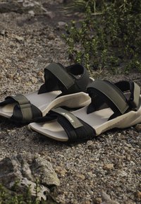 Czarne i beżowe sportowe sandały outdoor z regulowanymi paskami, spoczywające na żwirowym podłożu w pobliżu zielonych roślin.