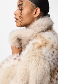 Beżowy płaszcz ze sztucznego futra z ciemnymi plamami, o puszystej fakturze i szerokim, miękkim kołnierzu. Widoczne detale na ramionach i rękawach.