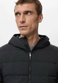 Zwarte doorgestikte jas met ritssluiting en kap. Heeft horizontale stiknaden en een grijze voering. Draagt over een grijze trui.