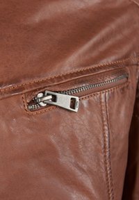 Braune Lederjacke mit einem Metallreißverschluss und rechteckigem Zug, strukturierte Oberfläche und detaillierte Nähte entlang der Säume.