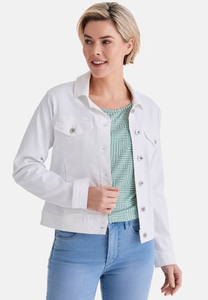 Frau mit kurzen blonden Haaren, die eine weiße Jacke mit Knöpfen über einem grünen gemusterten Hemd und hellblauen Jeans trägt, lächelnd und nach rechts schauend.