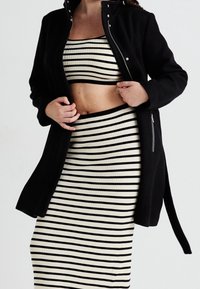 Černý vnější kabát s patentkami přes proužkovaný crop top a sukni v krémové a černé barvě, zdůrazňující přiléhavé svislé linie.