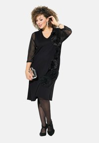 Schwarzes Kleid mit V-Ausschnitt, transparenten Dreiviertelärmel, verziert mit stofflichen Blumenakzenten auf einer Seite. Kombiniert mit einer metallischen Clutch und Absatzschuhen.