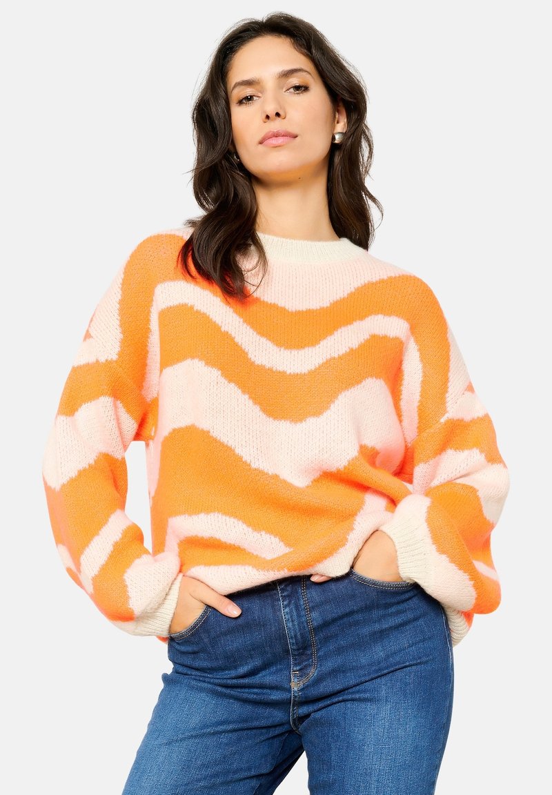 Gehaakte oversized trui in een oranje en roze zigzagpatroon, met ribgebreide cuffs en een ronde halslijn. Gecombineerd met blauwe spijkerbroeken.