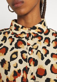 Blusa com colarinho em creme claro com manchas de leopardo pretas, laranjas e castanhas; apresenta fecho de botão e textura de tecido suave.