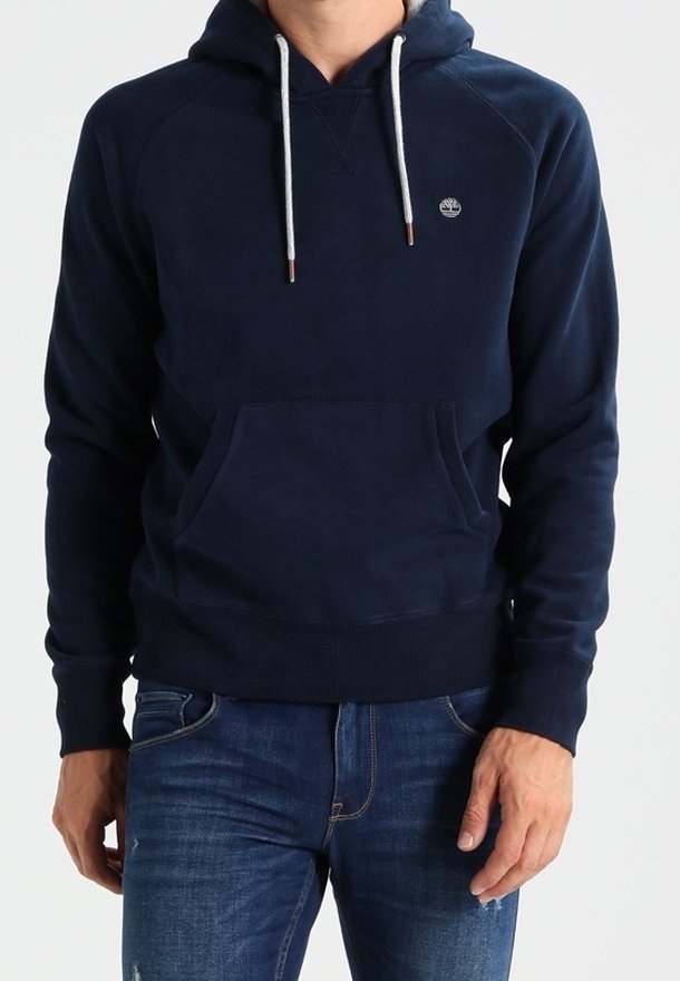 Homme portant un sweat à capuche bleu marine avec des cordons blancs et un petit logo sur la poitrine, associé à un jean bleu.