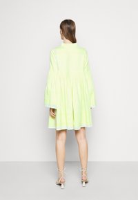 Neon žluté šaty s volánkovými rukávy, stáhnuté pod prsy a s rozšířenou sukní. Mají strukturovaný vzor a vysoký límec.