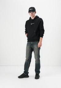 Schwarzer Hoodie mit Kängurutasche und Kordelzug, graue Jeans und schwarze Sneakers. Ergänzt durch eine schwarze Mütze mit weißem Text.