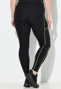 Leggings negros con costuras en acento verde, con un diseño ajustado y textura suave. Llevados con zapatos deportivos negros.