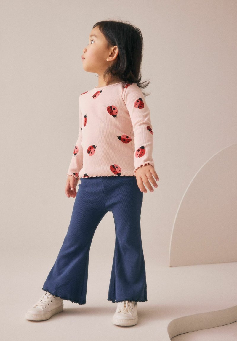 Jeune enfant en t-shirt rose à manches longues avec imprimé coccinelle, pantalon bleu évasé et baskets blanches regardant sur le côté sur un fond uni.