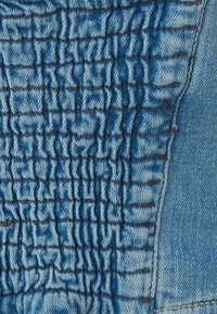Tessuto in denim blu chiaro caratterizzato da texture arricciata con linee di cucitura orizzontali. Le sezioni lisce contrastano con le aree plissettate.