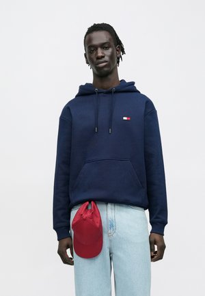 Uomo che indossa una felpa con cappuccio blu scuro e jeans azzurri con un cappellino rosso appeso alla cintura, in piedi davanti a uno sfondo chiaro e uniforme.