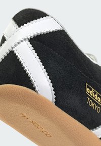 Melnas zamšādas sporta apavi ar baltām ādas akcentiem, zigrasta šuvēm un gumijas zoli. Zelta "adidas TOKYO" zīmols sānos.