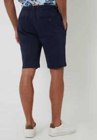 Shorts en coton bleu marine avec une taille élastique, deux poches arrière avec des boutons et des ourlets roulés. Portés avec des baskets blanches.