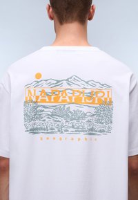 Vit bomulls-t-shirt med en grafisk design av berg och lövverk i ljusgrönt och orange, med texten "NAPAPIJRI geographic" nedanför.