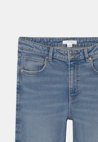 Modré džíny z denimu s předním zapínáním na knoflík, poutky na opasek a viditelným prošitím mincovní kapsy na bílém pozadí.