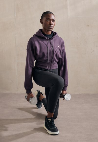 Purple cropped hoodie with drawstrings, black tank top, black leggings, and black sneakers. Holding silver dumbbells, kneeling on one knee.