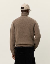 Καφέ, πλεκτό πουλόβερ με ψηλό, ριγέ γιακά και μακριά μανίκια; φορεμένο με μια μπεζ κάππα. Μαύρο παντελόνι από κοτλέ ορατό εν μέρει.