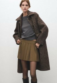 Manteau texturé marron en mélange de laine, superposé à un pull gris et une mini-jupe plissée kaki. Le mannequin porte des bottes noires montant jusqu'aux genoux.