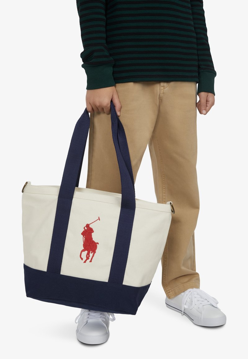 Torba na rame od krem platna s tamnoplavom podlogom i ručkama, s crvenim vezom logotipa igrača polom. Glatka tekstura, ležeran dizajn.