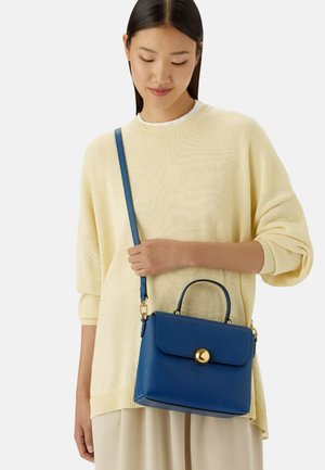 Femme en pull crème et pantalon, portant un sac à main en cuir bleu avec fermoir doré et bandoulière ajustable.
