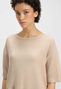 Beżowa, teksturowana bluzka z krótkim rękawem o szerokim dekolcie i luźnym kroju. Posiada delikatne prążkowanie oraz małą metkę marki z boku.