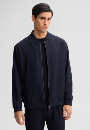 Uomo con capelli scuri che indossa una giacca blu navy con zip sopra una maglietta nera, in piedi davanti a uno sfondo grigio chiaro uniforme.