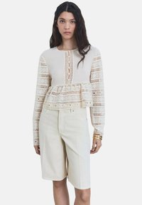 Top peplum crema con inserti in pizzo, maniche lunghe e bottoni a oblò; abbinato a pantaloni corti beige chiaro fino al ginocchio.