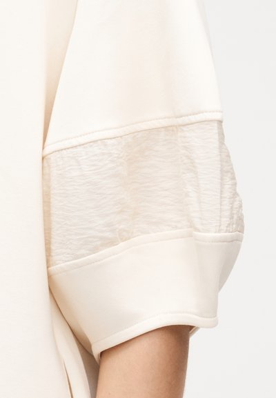 Κοντινό πλάνο μανσετίνας ένδυματος εκρού χρώματος με υφή από διάφανο τούλι ανάμεσα σε τμήματα συμπαγούς υφάσματος που τελειώνουν πάνω από τον αγκώνα.