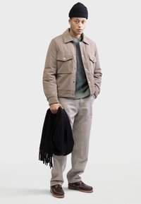 Bruine corduroy jas met twee borstzakken, groene gebreide trui, grijze broek, bruine schoenen en een zwarte sjaal. Model draagt een zwarte beanie.