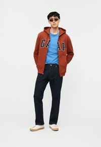 Νεαρός άνδρας με σκουρόχρωμο καφέ φούτερ GAP, μπλε μπλουζάκι, σκούρο τζιν, λευκά αθλητικά παπούτσια και γυαλιά ηλίου, που στέκεται μπροστά σε απλό φόντο.