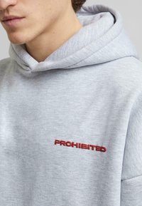Grå hettegenser laget av myk bomull, med rødt brodert tekst "PROHIBERT" på brystet, med en avslappet passform og teksturert stoff.