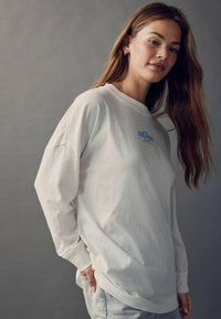 Tricou alb din bumbac cu mânecă lungă, având un mic logo albastru pe piept, croială lejeră și mansete ribbrate la încheieturi.