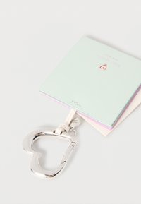 Τετράδιο σε μέντα πράσινο με ασημί καραμπίνερ σε σχήμα καρδιάς, διαθέτει τρεις χρωματιστές ενότητες και ανάγλυφο λογότυπο. Η επιφάνεια έχει λείο υφή.