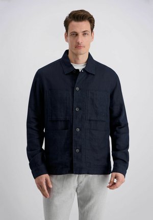 Man draagt een donkerblauwe jas met knopen en vier zakken aan de voorkant, over een wit shirt en lichtgrijze broek, staand tegen een effen achtergrond.