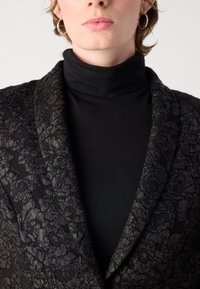 Zwarte blazer met een textuur van bloemmotieven, met een geklopte kraag. Draagt over een zwarte coltrui, die ingewikkelde details toont.
