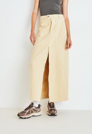 Beige, knee-length skirt with a front slit, made from cotton, featuring large pockets and a button closure. Paired with brown athletic sneakers.