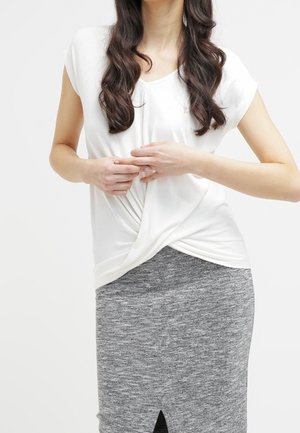 Femme aux longs cheveux bouclés portant un haut blanc noué à l'avant et une jupe crayon grise taille haute avec une fente à l'avant.